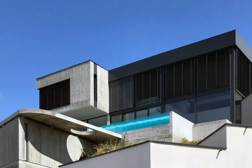 Maison contemporaine en béton et verre avec piscine surélevée sur une pente sous un ciel bleu clair - constructeur à lyon